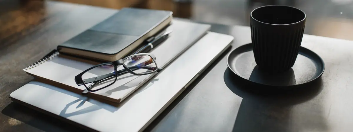Reading glasses on a stack of notebooks and a closed laptop beside a cup of black coffee