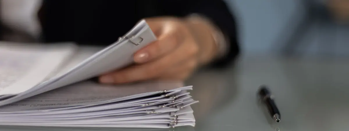 A hand sorting a thick stack of clipped documents on a glass desk beside a pen