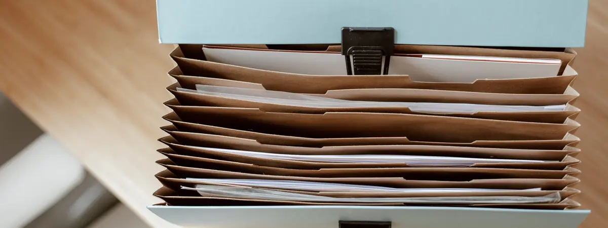 An open accordion file box with papers sorted into tabbed brown pockets on a wooden desk