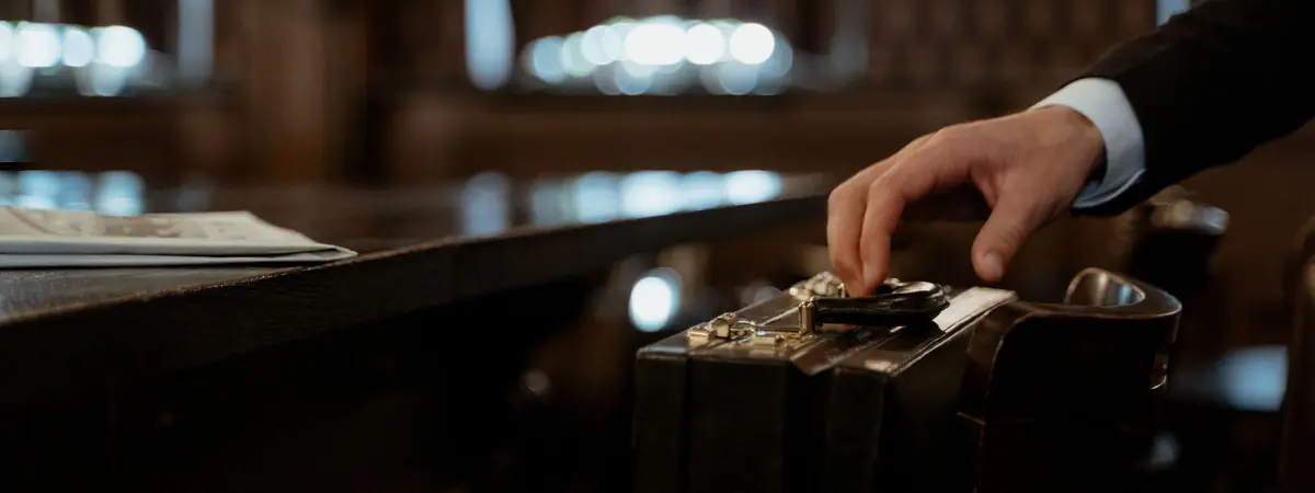 A hand in a suit sleeve resting on a black leather briefcase in a dim, wood-paneled room