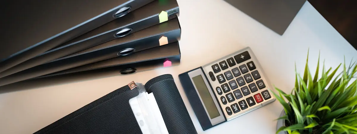 Black office binders marked with colored tab flags beside a desk calculator and a potted plant