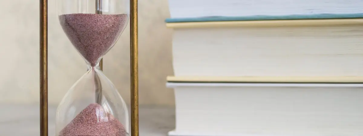 A brass hourglass with pink sand running through it, beside a stack of hardcover books