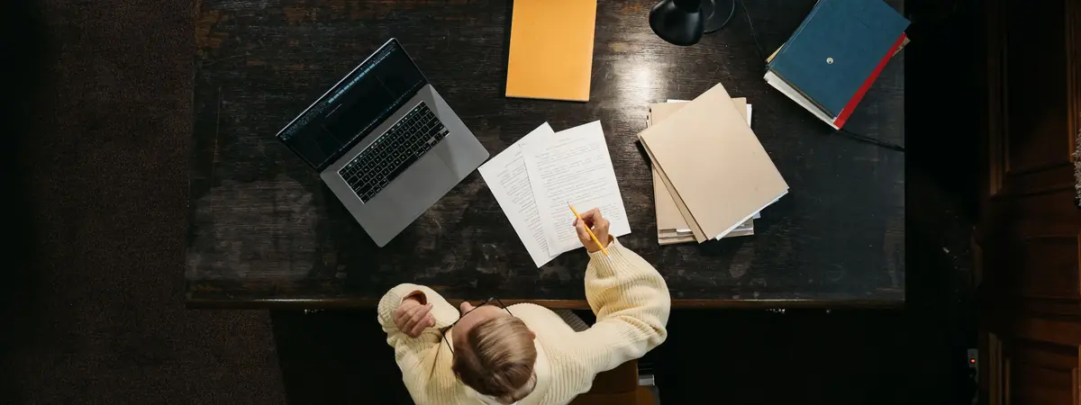 Seen from directly above, a person marks up printed pages with a pencil at a dark wooden desk
