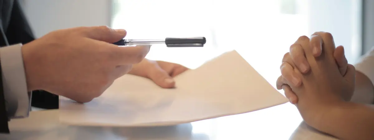 A professional in a suit hands a pen and a document across a table