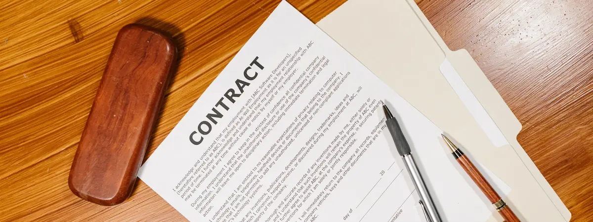 A printed contract with a signature line on a wooden desk beside two pens and a folder