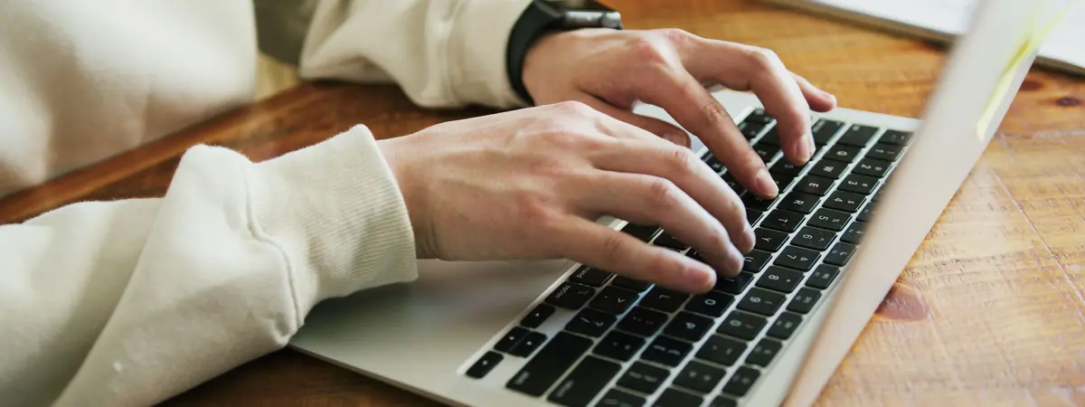 Close view of hands typing on a silver laptop keyboard at a wooden table