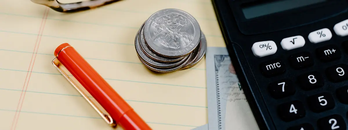 A calculator, a stack of quarters, a red pen and reading glasses on a yellow legal pad