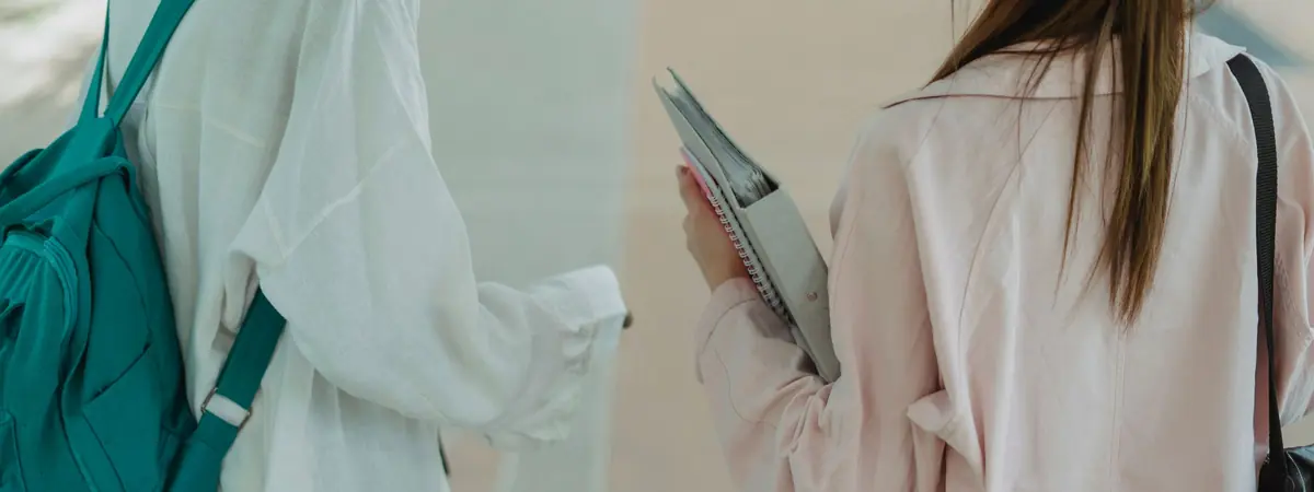 Two students seen from behind walk side by side, one carrying a binder and spiral notebooks