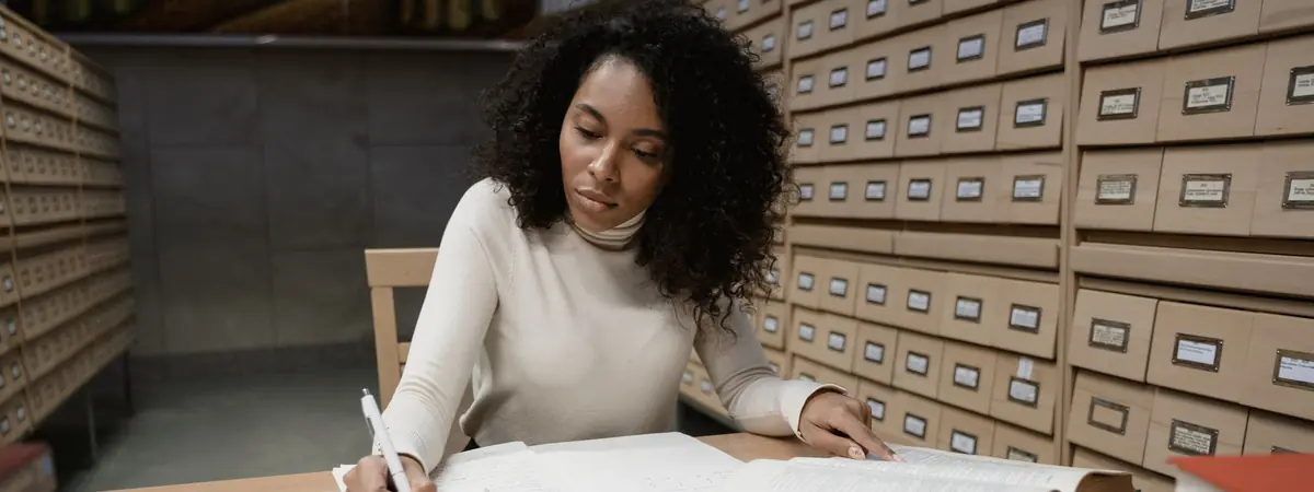 A woman in a cream sweater writes in a notebook beside open books at a library table