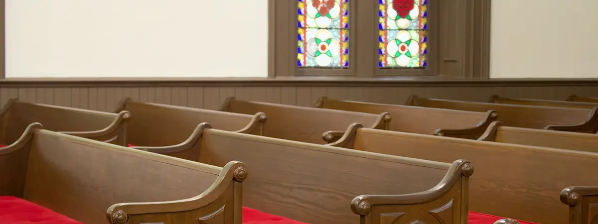 Rows of carved wooden pews with red cushions beneath two small stained glass windows on a cream wall