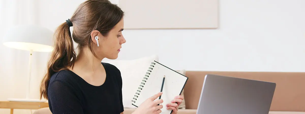 A woman with an earbud holds a spiral notebook toward her laptop in a bright living room
