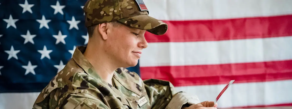 A soldier in a camouflage uniform and cap looks at a card in front of an American flag