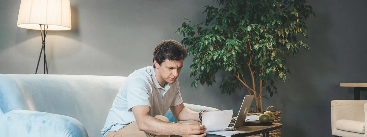 A man in a light blue polo sits on a sofa reading papers beside an open laptop