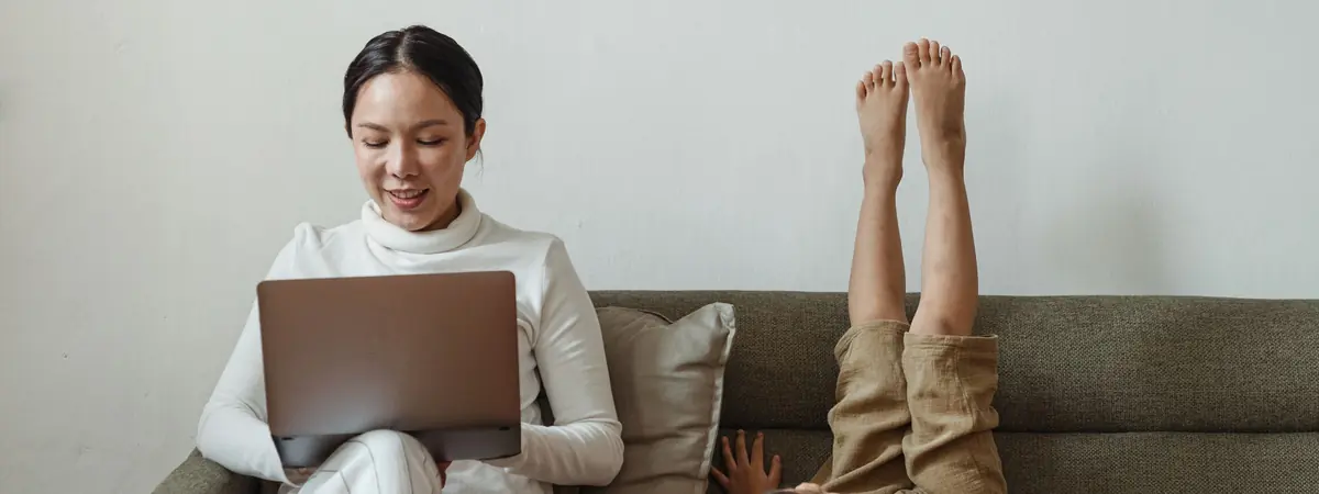 A woman smiles at a laptop on a green sofa as a child's feet stick up beside her