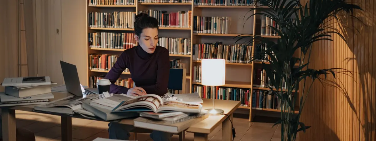 A woman studies at a wooden table beside a glowing lamp, with open books and shelves behind her
