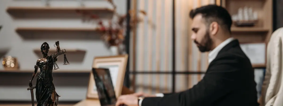 A man in a suit types on a laptop at a wooden desk beside a Lady Justice statue