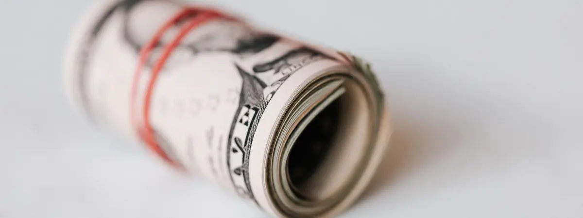 A roll of US dollar bills held by a red rubber band on a white tabletop